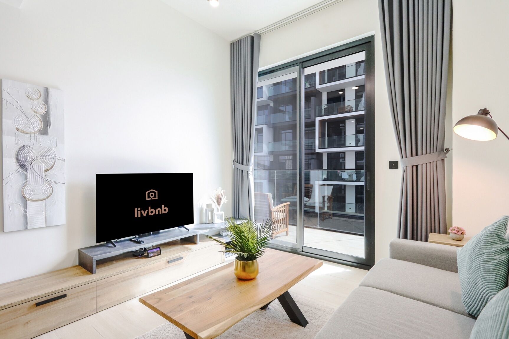 Un salon moderne doté d’un téléviseur à écran plat, d’une table basse en bois, d’un canapé et d’une grande porte-fenêtre coulissante donnant sur un balcon avec vue sur un immeuble d’habitation.