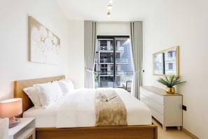 A modern bedroom with a bed, bedside table, lamp, window, and a view of the outside. - Chic 1BR in JVC with Balcony | Near Mall & Parks (Dubai)