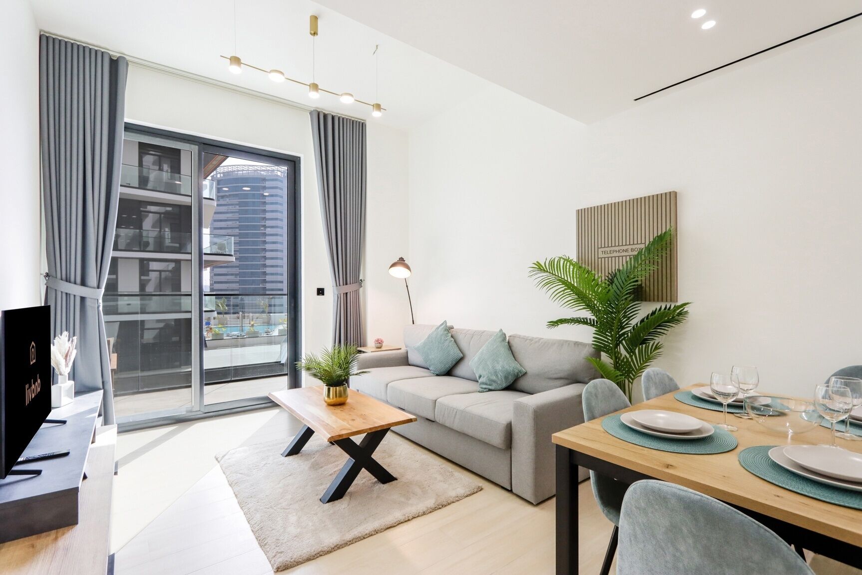 A modern living room with a sofa, a dining table set for two, a potted plant, and a view of a cityscape through large windows.