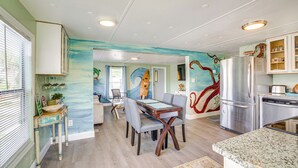 Un comedor con mesa y sillas, una cocina con refrigerador y armarios, y un mural en la pared.