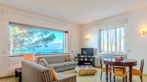 A living room with a sofa set, a TV, a dining table with chairs, and large windows with a view of the sea.