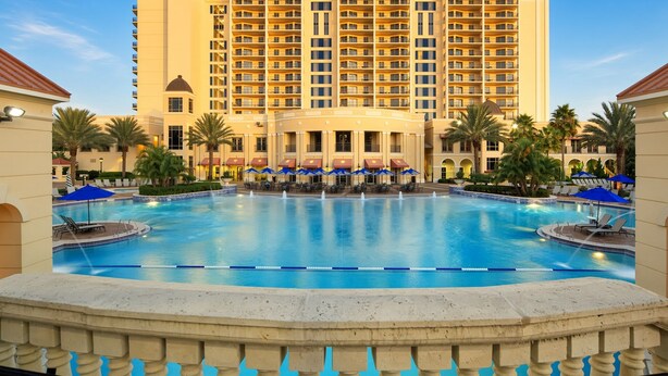 A large swimming pool with a fountain, surrounded by lounge chairs and umbrellas, with a hotel building in the background. - Orlando | Easy Disney Retreat | Pool + Parks (Orlando)