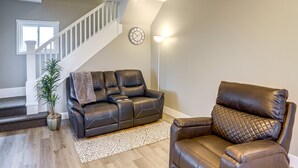 Een woonkamer met een bruine leren bank, een bruine leren fauteuil, een tapijt met een patroon, een potplant en een trap met witte leuning.