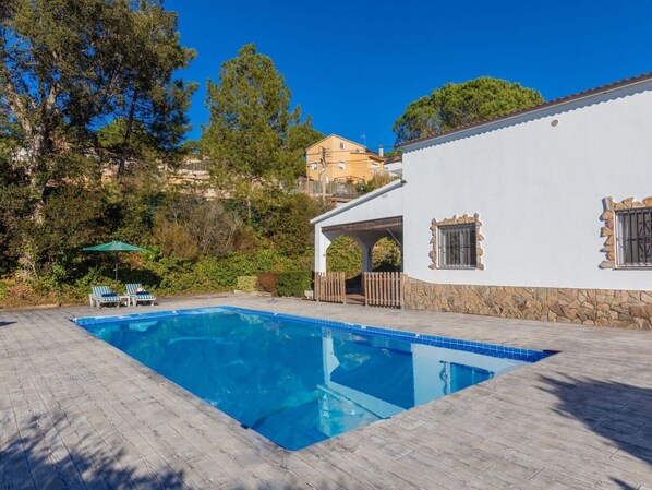 A swimming pool with a wooden deck, a white building with a stone base, and a green umbrella.