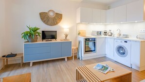 Eine kompakte Küche mit Mikrowelle, Toaster und Waschmaschine. Ein Fernseher steht auf einem Tisch und ein kleiner Tisch ist mit einer Zeitschrift bestückt.