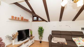 Ein Wohnzimmer mit Fernseher auf Holzfuß, einer Couch, einer Pflanze und einer Decke mit freiliegenden Balken.