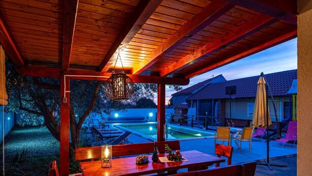 An outdoor patio with wooden tables and benches, a hanging light fixture, and a view of a swimming pool and houses in the background.