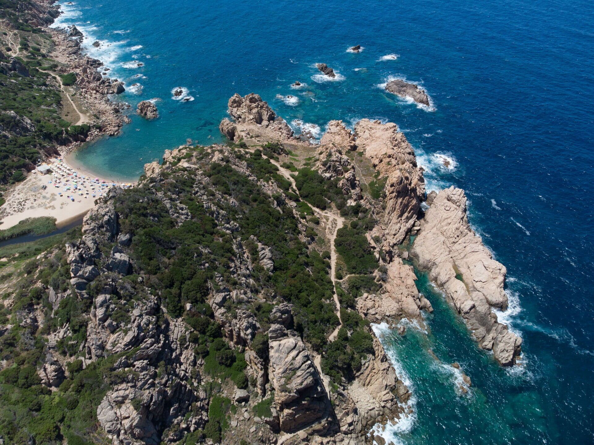 An aerial view of a coastal area with rocky terrain, a beach, and clear blue waters.