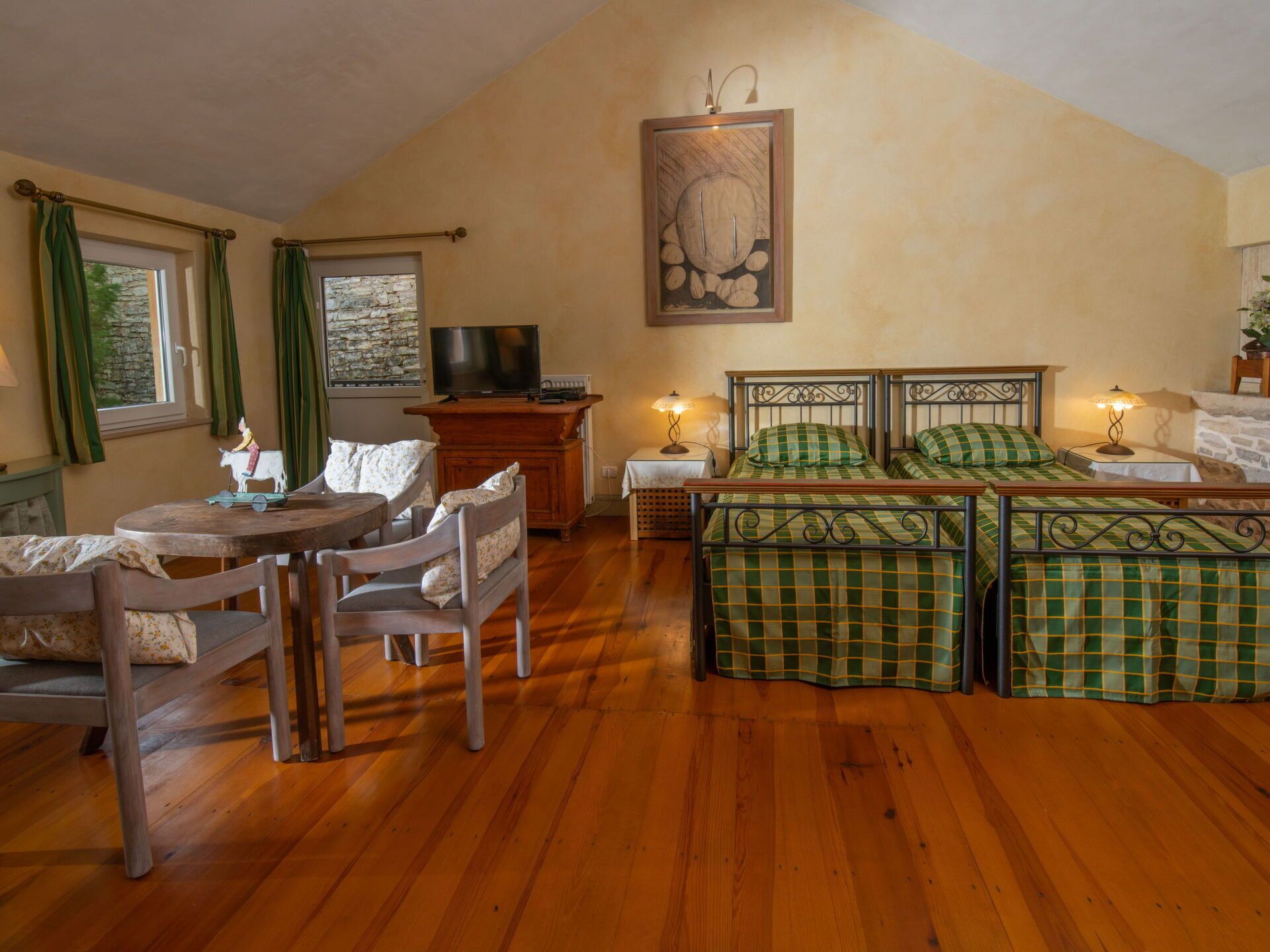 Une pièce chaleureuse avec un plancher en bois, une table à manger avec des chaises, un lit avec une literie à carreaux verts, un téléviseur et un tableau accroché au mur.