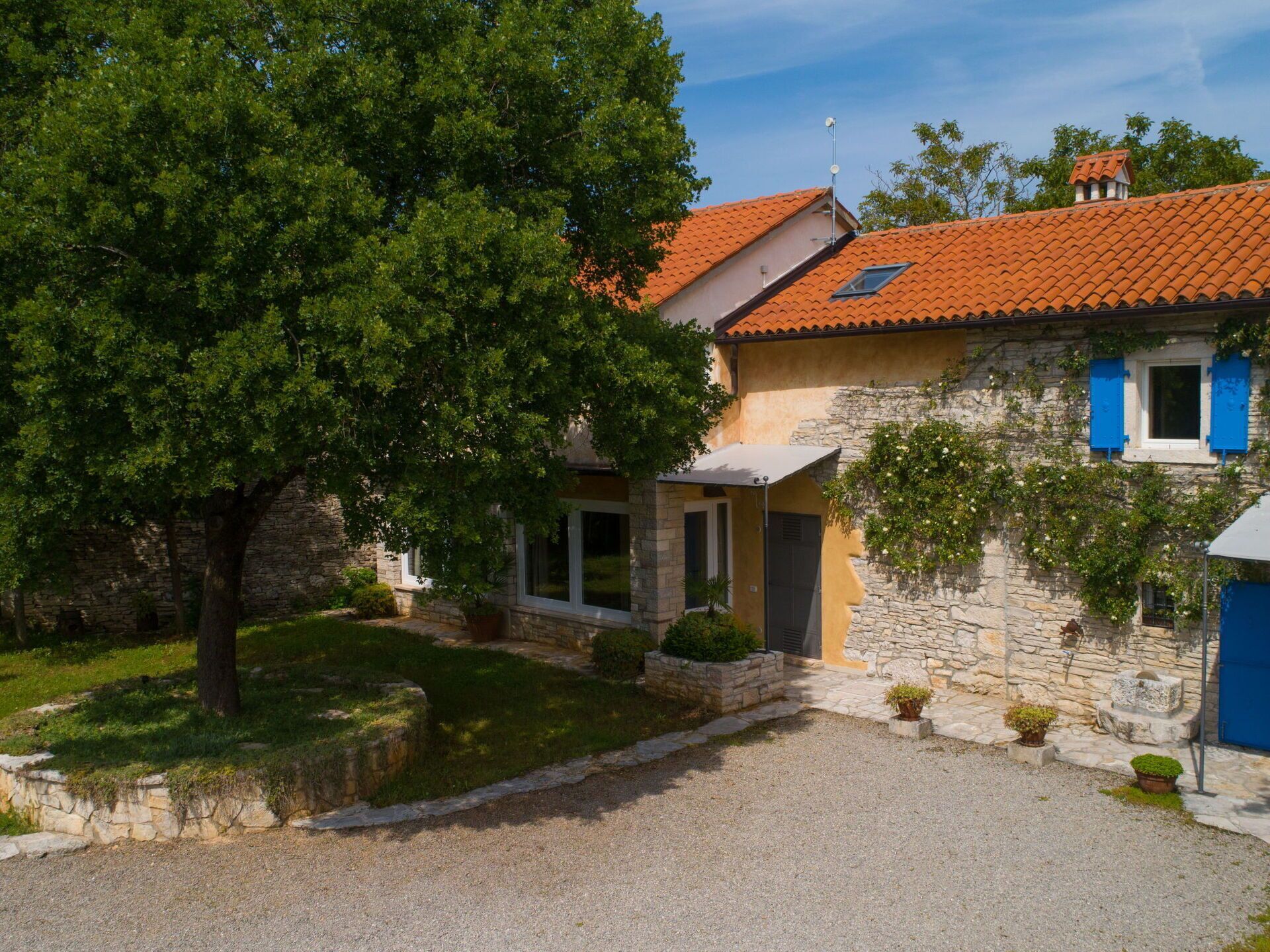 Une maison en pierre avec un toit de tuiles, un grand arbre dans le jardin et une allée gravillonnée.
