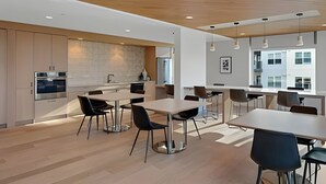 A modern kitchen with built-in appliances, a dining area with tables and chairs, and a view of a building outside. - 1BR+Den | Gym | Bellevue | Sophari Downtown D (Bellevue)