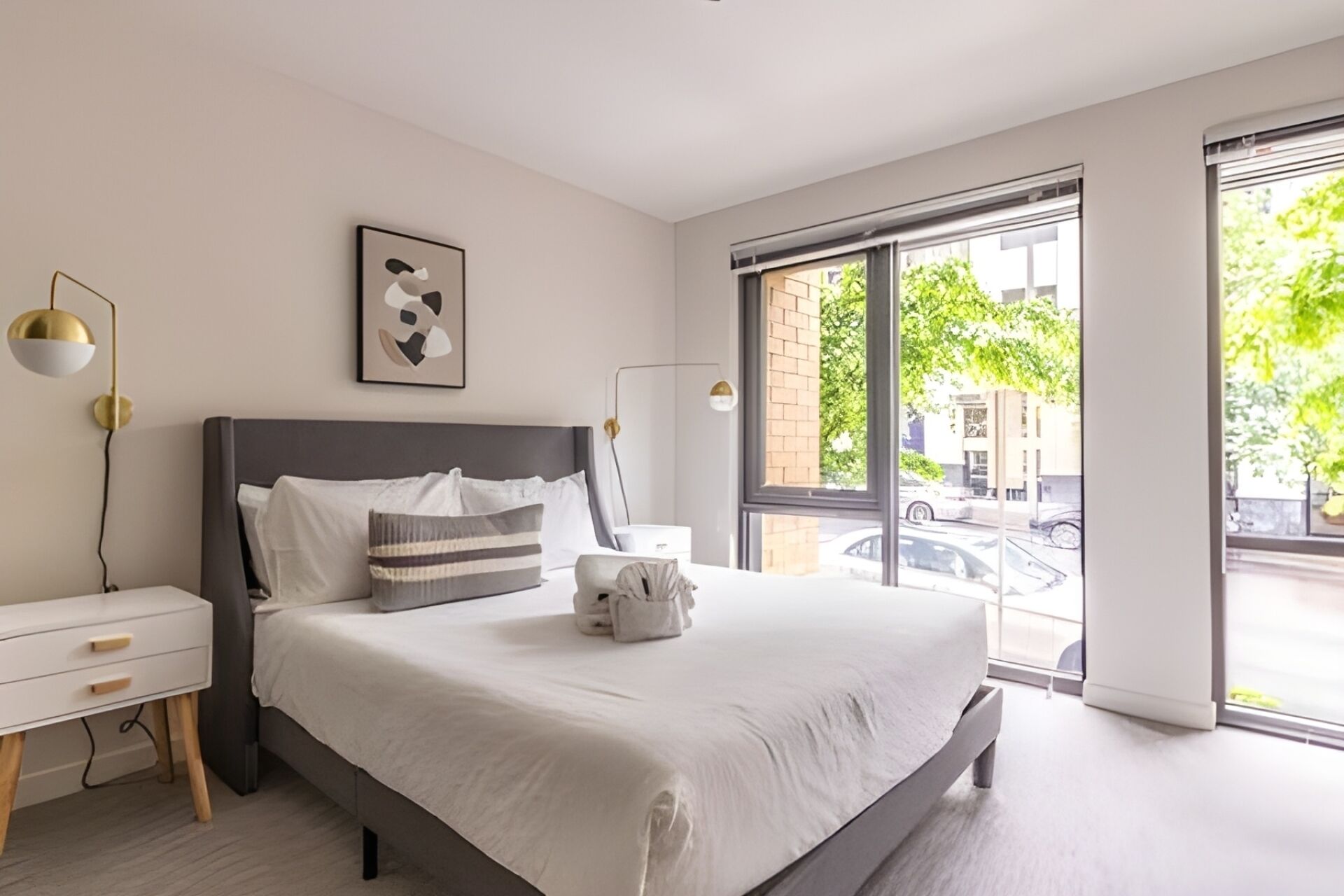 Ein modernes Schlafzimmer mit einem großen Bett, Nachttischen, einer Wandleuchte und einem Fenster mit Blick auf die Straße.