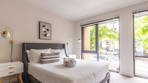 Une chambre moderne avec un grand lit, des tables de chevet, une lampe fixée au mur et une fenêtre donnant sur la rue.