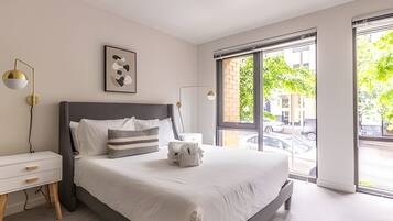 A modern bedroom with a large bed, bedside tables, a wall-mounted lamp, and a window with a view of the street.