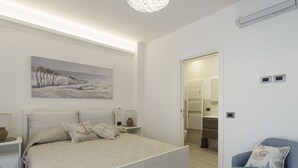 Ein modernes Schlafzimmer mit einem großen Bett, einem Nachttisch, einem Sessel, einem Gemälde und einem durch eine offene Tür einsehbaren Badezimmer.
