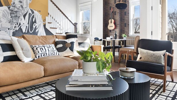 A modern living room with a brown leather sofa, a black coffee table with books, a guitar, and a dining area with a wooden table and chairs. - | Downtown Nash | Outlaw Theme | Large Groups | (Nashville)