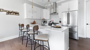 A modern kitchen with white cabinetry, a stainless steel refrigerator, and a bar area with stools. - | Downtown Nash | Outlaw Theme | Large Groups | (Nashville)