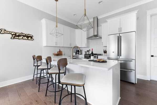 A modern kitchen with white cabinetry, a stainless steel refrigerator, and a bar area with stools. - | Downtown Nash | Outlaw Theme | Large Groups | (Nashville)