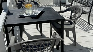 Une terrasse extérieure avec une table noire, des chaises blanches et un plateau contenant des verres et une carafe.