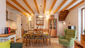 A cozy living room with a dining area, wooden beams on the ceiling, a green armchair, and a kitchen with modern appliances.