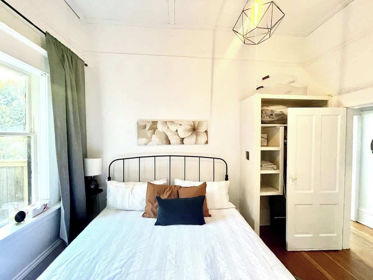 Ein Schlafzimmer mit einem Bett, einem Nachttisch, einer Lampe, einem Fenster mit Vorhängen, einem Wandbild und einem Kleiderschrank.