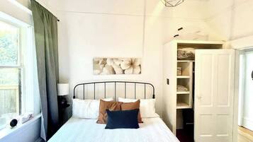 Ein Schlafzimmer mit einem Bett, einem Nachttisch, einer Lampe, einem Fenster mit Vorhängen, einem Wandbild und einem Kleiderschrank.