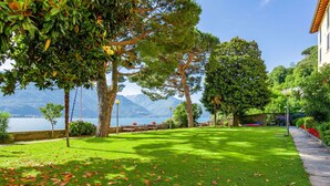 A well-maintained lawn with scattered fallen leaves, a stone pathway, and a bench area overlooking a lake with mountains in the background. - Lovely apartment for 4 guests with WIFI and TV (Vignola)