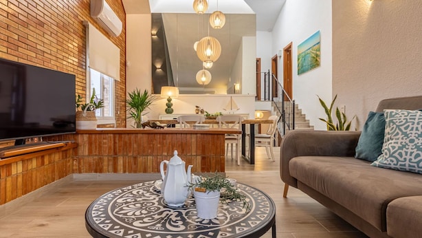 A modern living room with a flat-screen TV, a coffee table with a decorative centerpiece, a sofa with patterned cushions, and a staircase leading to an upper level.