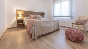 A bedroom with a wooden headboard, a bed with pink bedding, a bedside table with a lamp, a window, and a wooden chair.