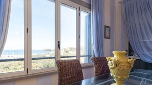A room with a glass table, wicker chairs, a yellow vase, and a view of the sea through large windows.