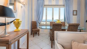 A room with striped wallpaper, a large window with curtains, a wicker chair, a glass table, a sofa, and a lamp.