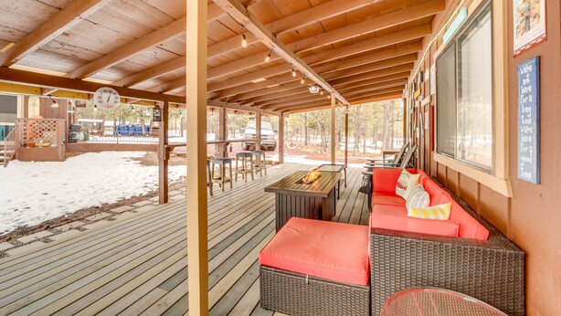 A covered outdoor seating area with a wooden deck, a table with a fire feature, and a red cushioned bench. - 2 Fire Pits & Covered Deck: Cabin in Happy Jack! (Happy Jack)