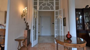 Un couloir avec une porte vitrée donnant sur une pièce, une table ronde ornée d’un vase et un meuble contenant des objets décoratifs.