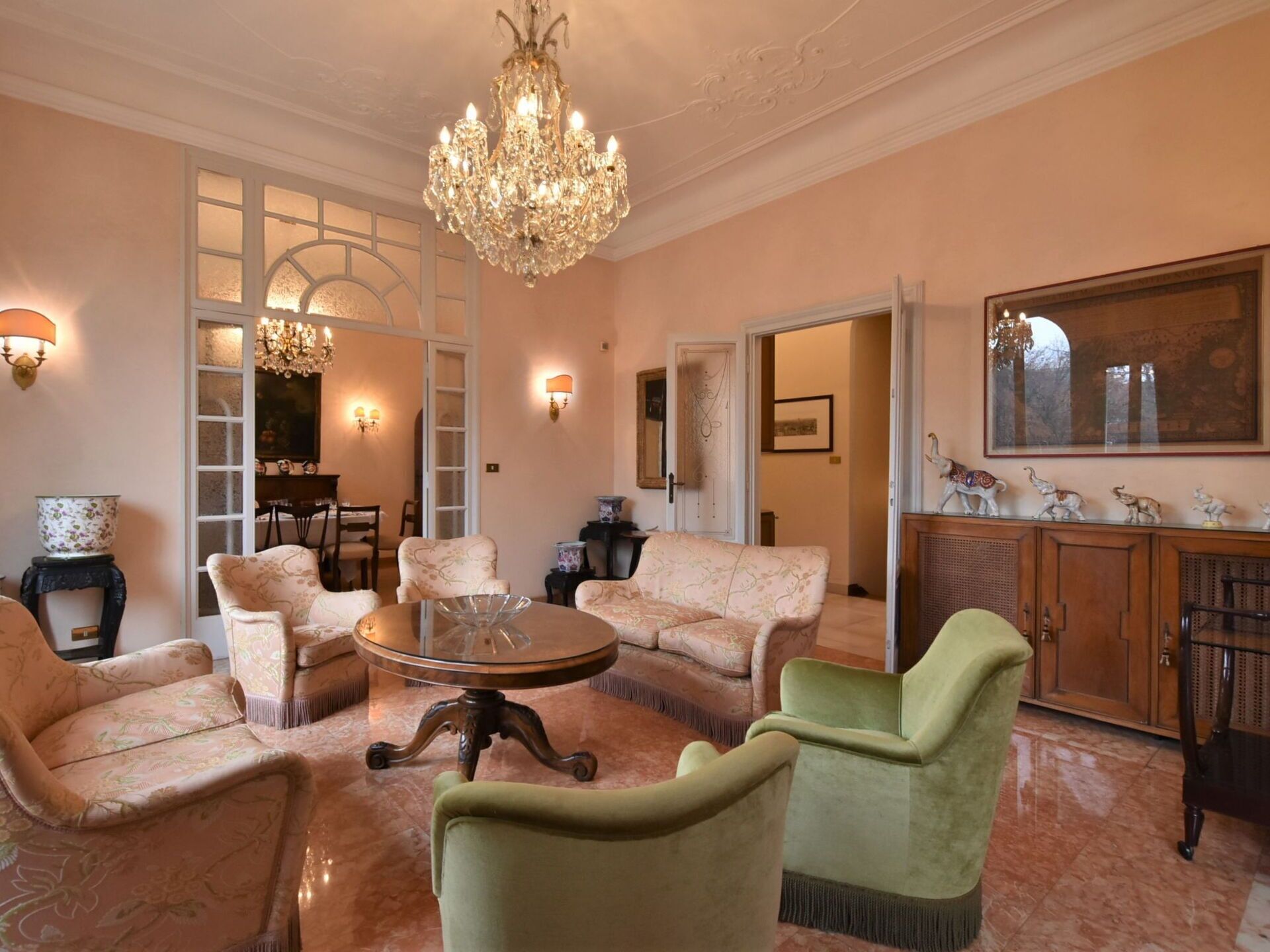 A spacious living room with a round coffee table, floral-patterned sofas, a chandelier, and a large mirror above a wooden cabinet.