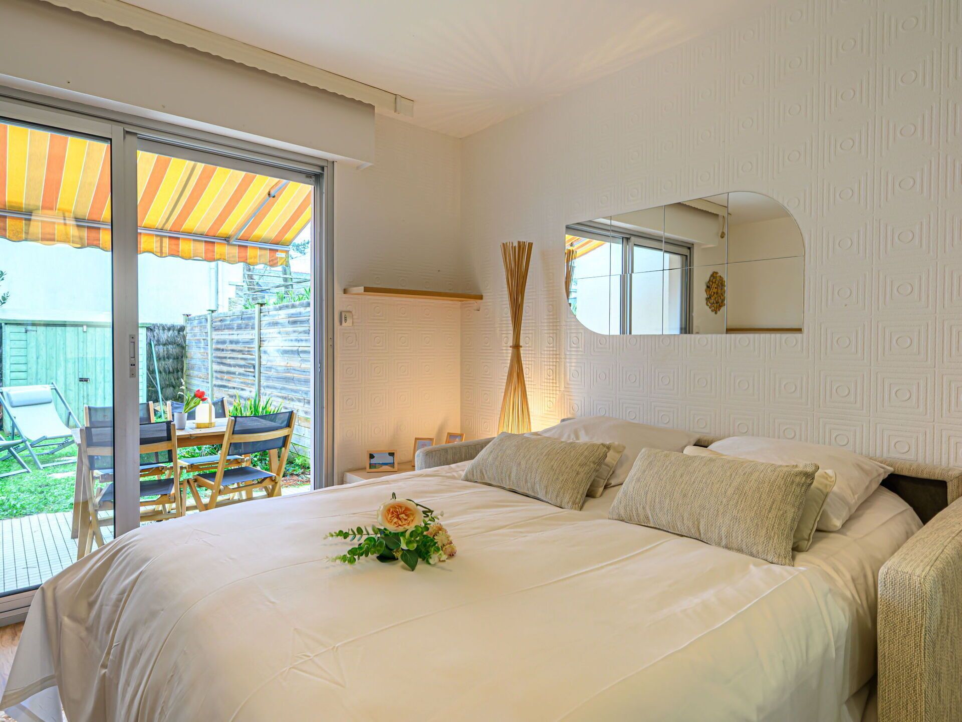 Ein Schlafzimmer mit einem großen Bett, einem kleinen Tisch und einem Sessel. Draußen befindet sich ein Fenster mit einem gestreiftem Sonnensegel.