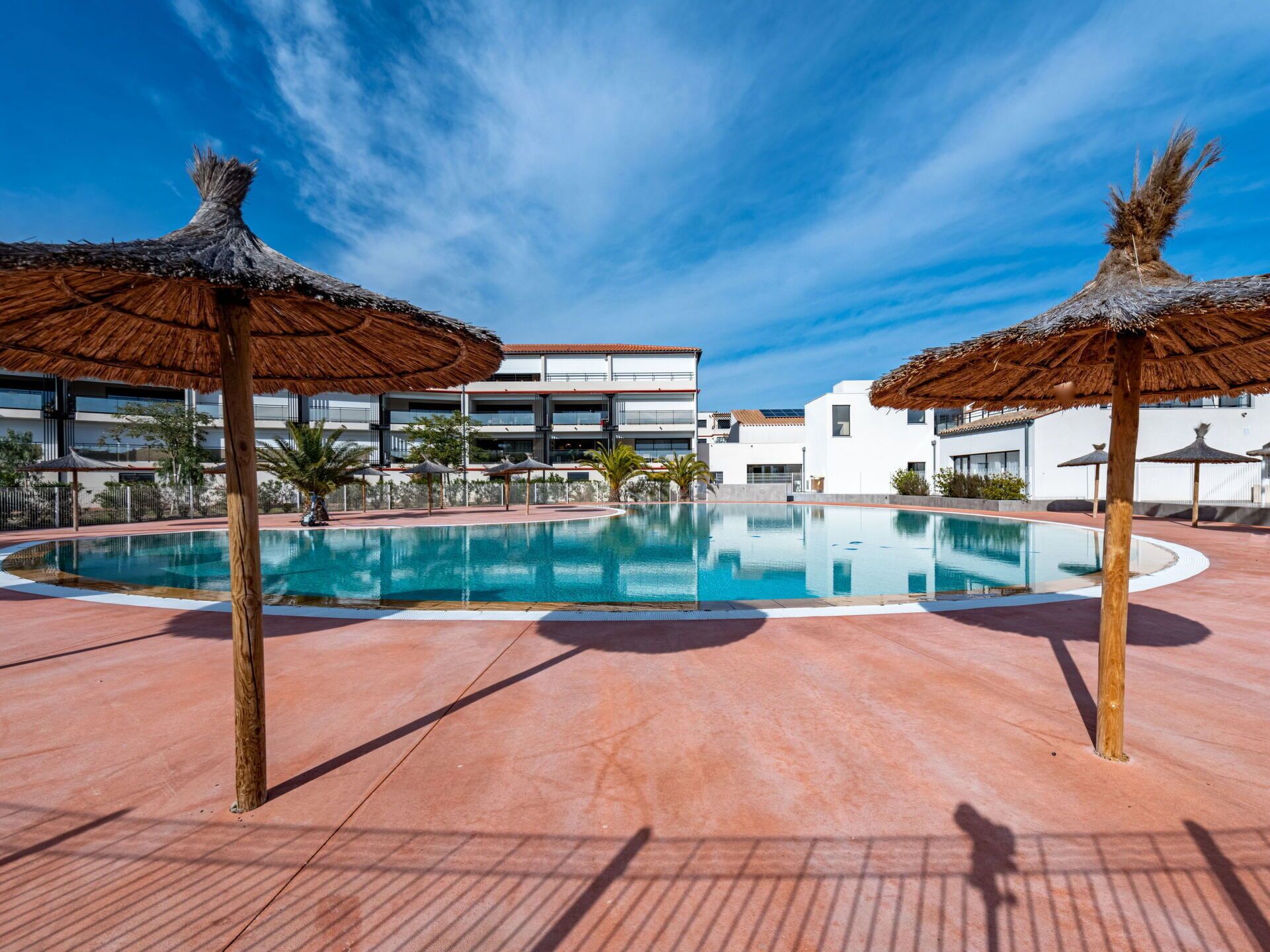 A swimming pool with thatched umbrellas, a clear blue sky, and modern buildings in the background.