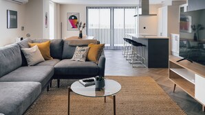 A modern living room with a grey sofa, a round glass coffee table, and a kitchen area with a bar and stools. - Luxe Penthouse Retreat: With Free Parking! (London)