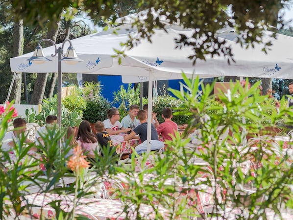 An outdoor dining area with tables and chairs under white canopies, surrounded by greenery.