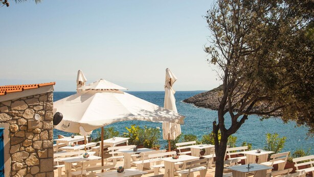 A seaside patio with white tables and umbrellas, stone walls, and a view of the sea. - Lovely apartment in villa for 4 guests with A/C, TV, terrace and pets allowed (Mali Lošinj)