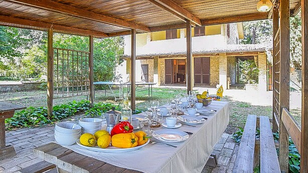 Un espace de restauration en plein air avec une longue table dressée pour un repas, entouré d’une pergola en bois et d’un sol en pierre.