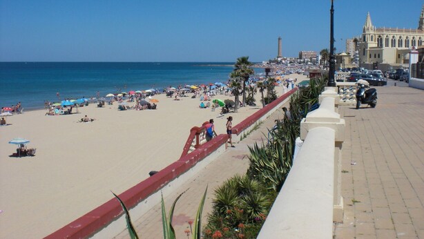 Ein Strand mit gepflastertem Weg, Palmen und einem historischen Gebäude im Hintergrund.