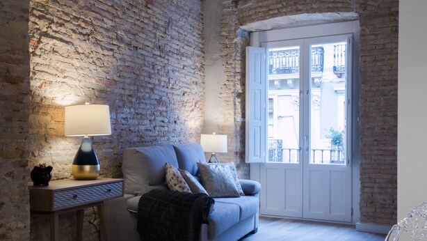 Ein gemütliches Wohnzimmer mit Backsteinwand, einer Couch, zwei Lampen und Blick durch eine Tür.