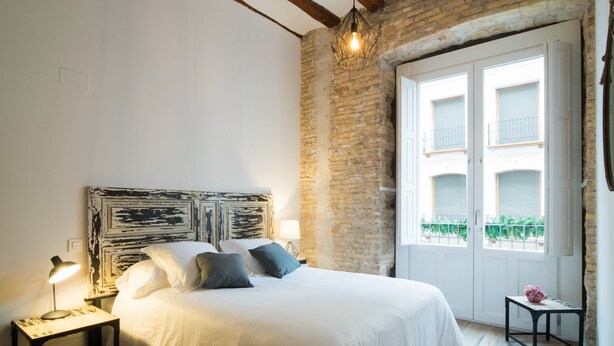 Une chambre avec un grand lit, une table de chevet avec une lampe, un mur en briques et une fenêtre donnant sur un balcon.