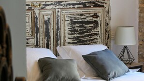 Un lit avec du linge de lit blanc et deux oreillers gris, un mur rustique dont la peinture s’écaille, et une lampe moderne posée sur une table de chevet en bois.