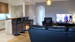A modern living room with a black leather sofa, a dining area with a bar, and a TV displaying a show. - Lovely apartment for 6 people with WIFI, pool, TV, balcony and panoramic view (Le Petit-saconnex)