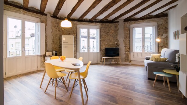 Amplia sala de estar con techo de madera, una mesa de comedor con sillas amarillas, un sofá gris y un televisor sobre un mueble de madera.