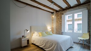 Un dormitorio con piso de madera, una cama con ropa de cama blanca, una mesita de noche con lámpara, una silla y una ventana con vista a edificios.