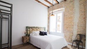 A bedroom with a wooden floor, a bed with white bedding, a nightstand, a chair, and a window with a view of the outside. - PPRINCIPAL B - P B - APARTAMENTOS ALMADA - Plaza del Pilar (Zaragoza)