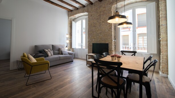 A modern living room with a sofa, yellow armchair, dining table, and chairs. There is a TV on the wall and a brick wall. - PPRINCIPAL B - P B - APARTAMENTOS ALMADA - Plaza del Pilar (Zaragoza)