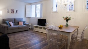 A modern living room with a grey sofa, a wooden dining table, clear chairs, a TV on a white stand, and a chandelier.